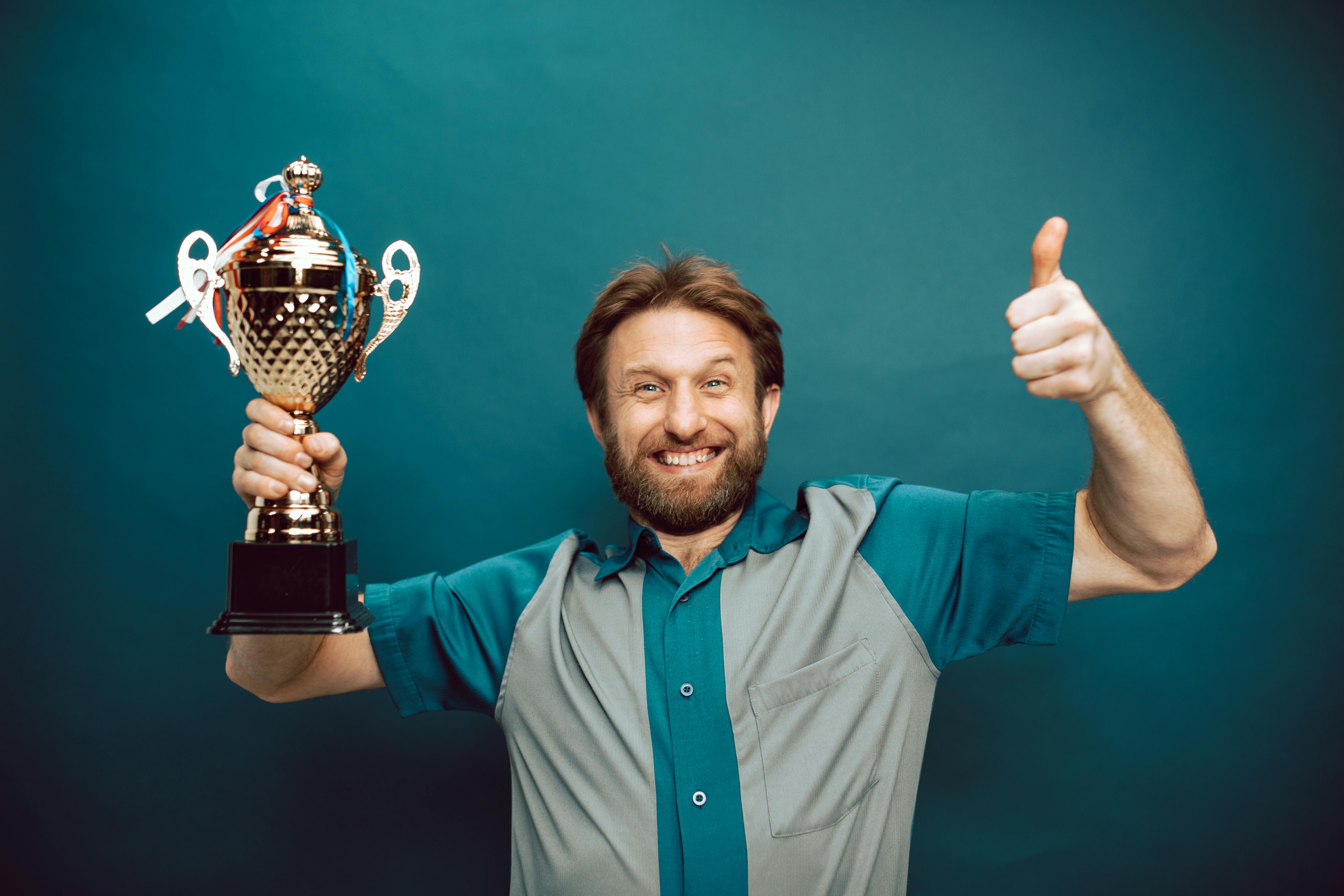 a bearded man grins ear to ear, likely because he's holding the trophy from a competition he won